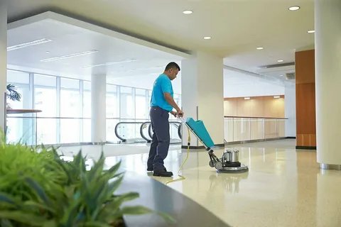 A janitor in a blue shirt and gray pants is using a large floor-polishing machine to clean the shiny, speckled floor of a modern commercial building lobby.